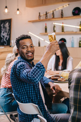 cheerful african american man looking at camera while holding glass of beer near multicultural friends