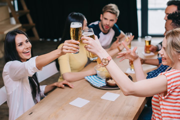 happy young woman clinking glasses of light beer while sitting in pub with multicultural friends