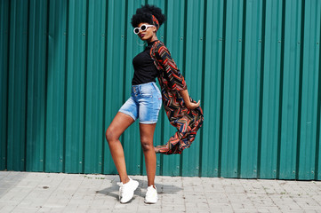 African woman with afro hair, in jeans shorts and white sunglasses posed against green steel wall.