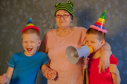 elderly woman in pink T-shirt and glasses holding megaphone. Next to grandmother, grandchildren in festive clothes in clown hats and patch on nose. Kids love grandma and have good time with granny