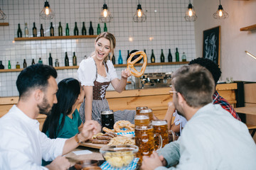 cheerful waitress in traditional german costume holding pretzel while standing near friends in pub
