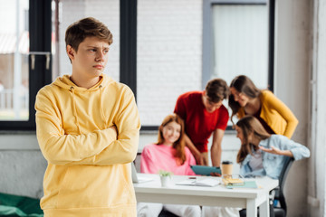 Fototapeta premium sad boy in yellow hoodie with crossed arms and laughing teenagers at desk