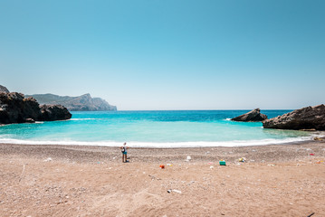 Fototapeta na wymiar Olympos, Karpathos/ Greece - 07 09 2019: volunteer woman starting beach cleanup and walking on a beautiful beach full of plastic rubbish litter in background is the blue Aegean Sea holiday destination