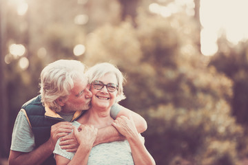 Couple of happy and cheerful people caucasian senior hug with love and smile with defocused green forest in background - outdoor leisure activity for retired matures- happiness for old