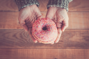 Vertical close up top view of sweet pink donuts holding by adult woman hands - wooden trendy hipster table in background - concept of breakfast and uhealthy food lifestyle
