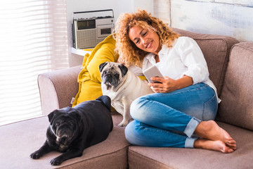 Cheerful woman at home sit down on the sofa with her two best friends dog pug near her - real lifestyle people with animals