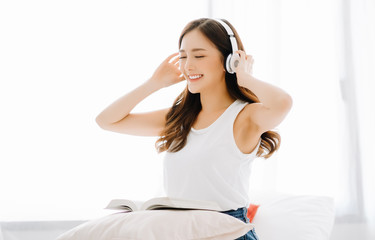 Portrait of young Asian woman smiling friendly listening music in headphones and dancing in white bedroom.Concept woman relax lifestyle.