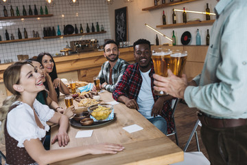 partial view of man holding glasses of light beer near multicultural friends in pub