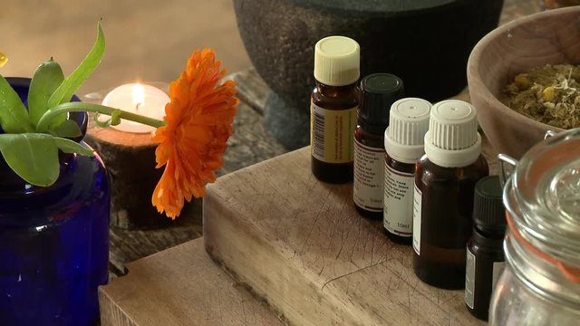 Steady, medium close up shot of multiple bottles of oils next to a candle and an orange flower.