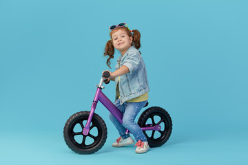Little girl learns to keep balance while riding a bicycle. Stylish child in sunglasses  isolated on blue background