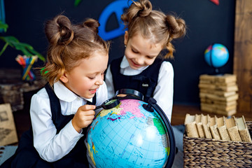 Little twin girls in school uniform using magnifying glass to examine globe while sitting on floor during geography lesson