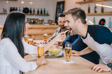 young woman feeding cheerful man with french fries while sitting in pub with friends