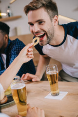 partial view of woman feeding cheerful man with fried onion ring while sitting in pub with multicultural friends