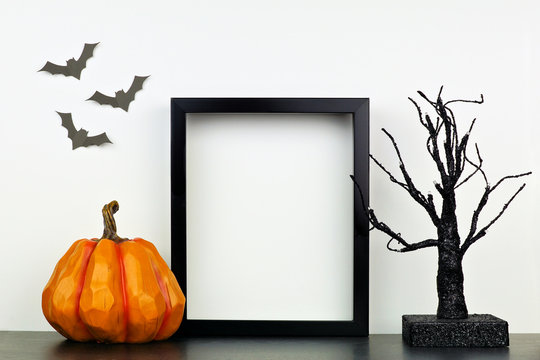 Mock up black frame with pumpkin and spooky tree decor on a shelf or desk. Halloween concept. Portrait frame against a white wall with bats.