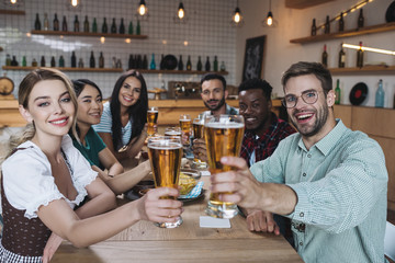 cheerful multicultural friends holding glasses of light beer and smiling at camera