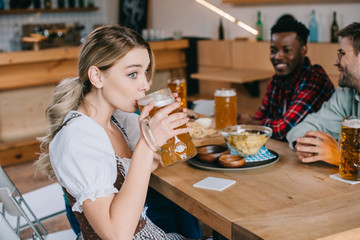 selective focus of young woman in traditional german costume drinking beer near cheerful multicultural friends