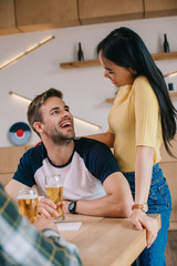 young asian woman standing near friend sitting with glass of beer in pub