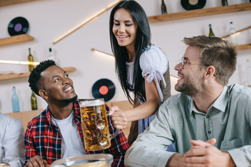 beautiful waitress in traditional german costume serving beer for multicultural friends in pub