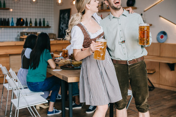 cropped view of cheerful man and woman in traditional german costumes holding mugs of beer
