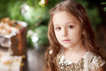 Portrait of a cute long-haired little girl in dress on background of Christmas tree and lights. Christmas and New Year celebration concept. Winter holidays. Close up