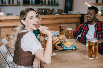 cheerful young woman eating fried sausage while celebrating octoberfest with multicutiral friends