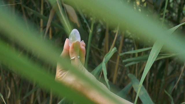 Female hand with french manicure holding transparent violet amethyst yoni egg for vumfit, imbuilding or meditation. Crystal quartz egg in hands on green background outdoors.