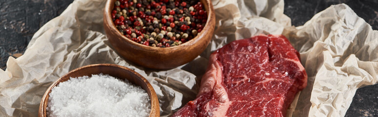 panoramic shot of raw beef fillet near wooden bowls with salt and peppercorns on parchment paper