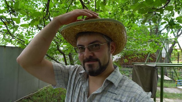 A man in glasses with a beard and a straw hat. Speaks and gestures with his hands. Portrait to the waist. One man. Against the background of green plants. The rustic atmosphere.