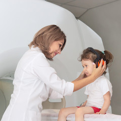 The child sits on a drawer of an MRI machine. Nearby A father and a female doctor are preparing a little girl for magnetic resonance imaging in a hospital. Nurse putting headphones on baby