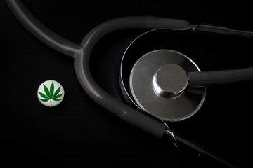 medical stethoscope and one white tablet with a marijuana leaf pin on a black background, medical cannabis concept, top view, close-up, copy space