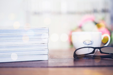Stack of magazine  and a cup of coffee, eye glasses on wooden table in dark room with bokeh light effected