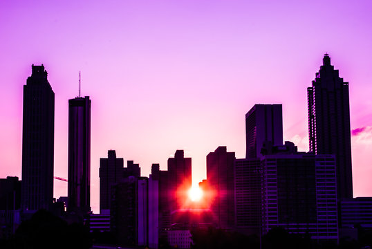 The sun sets on a city skyline which creates a stunning silhouette effect underneath a colorful sky. The buildings boast strong shapes against the soft color of the sky
