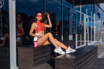 Stylish happy young brunette woman wearing white shorts and sneakers holding pink cup of coffee to go sitting next to coffee shop.