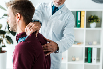 cropped view of chiropractor in white coat touching back of patient in clinic