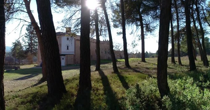 Dron vuela cerca del suelo sorteando arboles de un bosque junto a ermita abandonada en borque mediterraneo