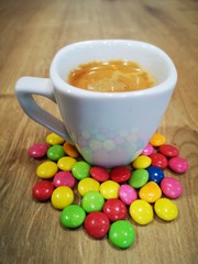 White cup with espresso coffee with colored chocolate candies on wooden table
