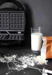 Cook at home. Waffle iron, flour, a glass of milk and flour on a dark background. A slice of the kitchen while cooking
