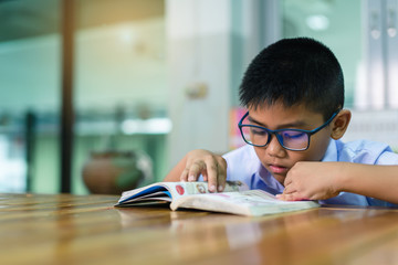 A cute Asian elementary school boy wearing blue glasses in a white school uniform is sitting, enjoying , reading comics in the library.