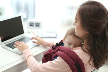 Young woman with baby in ergo carrier using laptop indoors