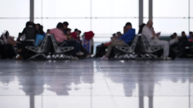 Airport lounge, blurred view of passengers sitting and waiting for boarding, slow motion shot. Bright terminal window on background, camera focused on floor at foreground