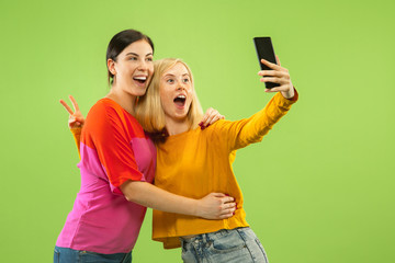 Portrait of pretty charming girls in casual outfits isolated on green studio background. Girlfriends or lesbians making selfie. Concept of LGBT, equality, human emotions, love, relation.