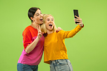 Portrait of pretty charming girls in casual outfits isolated on green studio background. Girlfriends or lesbians making selfie. Concept of LGBT, equality, human emotions, love, relation.