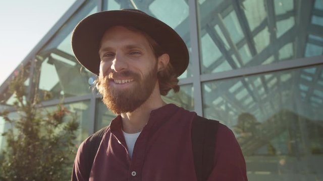 Close up of young red-haired Caucasian man with beard standing against glass wall outdoors, looking at camera and laughing