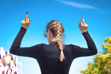 A young slender woman shows the middle finger with two hands, turning her back, the blue sky in the...