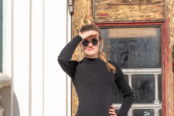 Young attractive woman in sunglasses covers her eyes with her hand from a very bright sun. The old wooden door of a large building in the background,