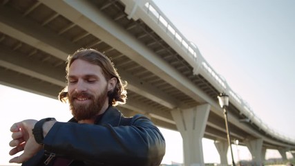 Low angle view of cheerful bearded red-haired man walking, checking time on his smart watch, looking at camera and then looking aside
