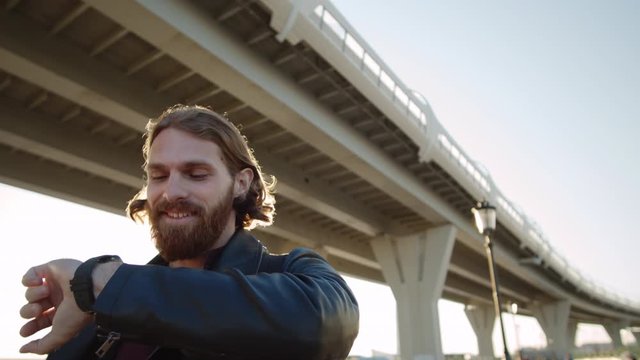 Low angle view of cheerful bearded red-haired man walking, checking time on his smart watch, looking at camera and then looking aside