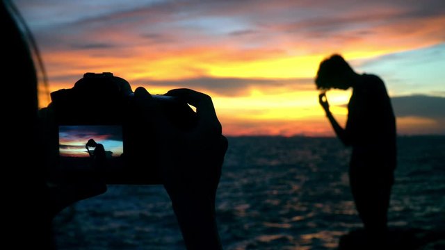 4K Wide shot of silhouette young adult romantic couple woman taking photo with digital camera of man boyfriend standing at rocky coastline  at sunset with twilight sky in summer travel vacations.