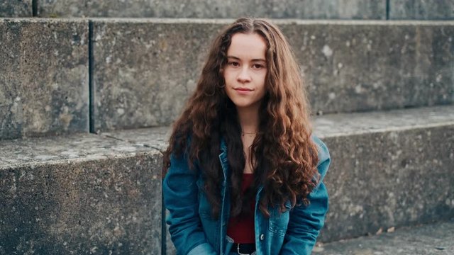 Portrait shot f the cute and stylish teen Caucasian girl with long hair sitting on the stoned steps outside, looking at the side and turning her face to the camera with a smile.