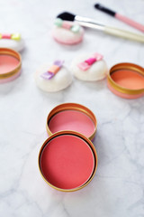 Small round blusher pots with different shades of pink and cute powder puffs with ribbons and makeup brushes in background